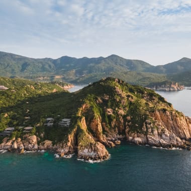 Aerial view of a rugged coastline with lush green hills and clear blue waters, with mountains in the background.