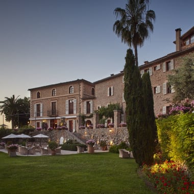 Ein großes historisches Hotel mit Steinwänden und mehreren Fenstern, umgeben von einem gepflegten Garten mit Bäumen, Blumen und Sitzbereichen im Abendlicht.