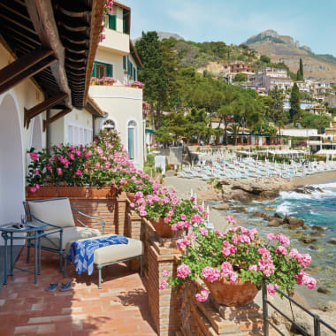 Ein gemütlicher Balkon mit pinken Blumen in Terrakottatöpfen, der auf die felsige Küste und bunte Gebäude in einem Küstendorf in Italien blickt.