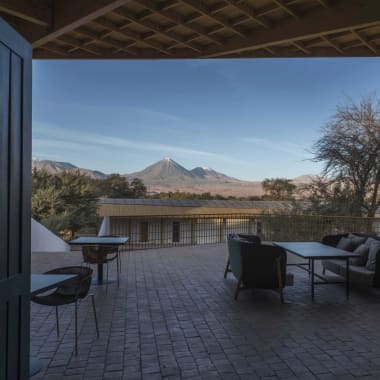 Ein Lounge-Bereich mit Sitzgelegenheiten im Freien blickt auf eine Wüstenlandschaft mit Bergen im Hintergrund, gesehen von einer schattigen Terrasse.
