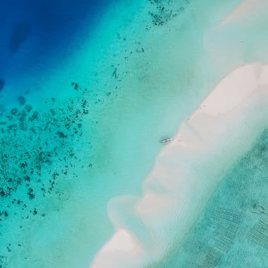 Luftaufnahme eines weißen Sandstrands mit klarem türkisfarbenem Wasser und dunklen Stellen von Korallen oder Felsen.