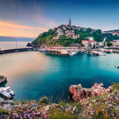 Lebendige Küstenansicht des Dorfes Vrbnik auf der kroatischen Insel Krk mit historischen Gebäuden auf einem Hügel mit Blick auf die Adria und Boote im Hafen bei Sonnenuntergang.