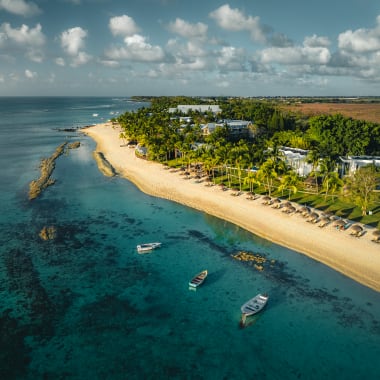 Luftaufnahme eines tropischen Strandresorts mit goldenem Sand, Palmen, mehreren Booten nahe der Küste und Sonnenschirmen entlang des Strandes unter teilweise bewölktem Himmel.