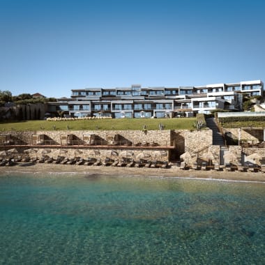 Modernes mehrstöckiges Hotelgebäude mit Blick auf ein klares türkisfarbenes Meer, mit Liegestühlen am Strand und Steinterrassen.