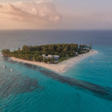 A small, lush island with trees and a few buildings surrounded by clear blue ocean, viewed from above at sunset.