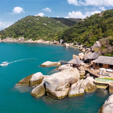 A serene view of a tropical bay with large rocks, lush green hills, and traditional wooden huts along the shoreline, with a boat cruising in the blue waters.