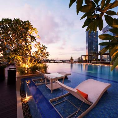 Luxuriöser Dachpool mit Blick auf die Skyline Singapurs bei Dämmerung, mit Poolliegen und üppigem Grün.