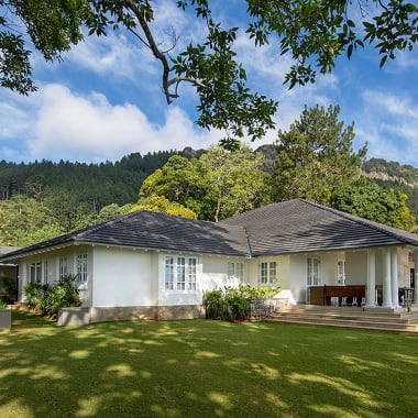 A large white house with a porch and steps, set in a lush green yard with trees and benches, against a backdrop of hills and blue sky with clouds.
