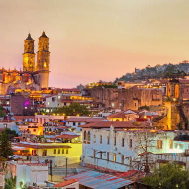 Panoramablick auf Taxco mit beleuchteten historischen Gebäuden und Kirchtürmen bei Sonnenuntergang.