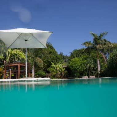 Ein Swimmingpool mit türkisfarbenem Wasser vor üppigem Grün und tropischen Bäumen unter einem blauen Himmel, mit einem weißen Sonnenschirm und Gartemöbeln in der Nähe.