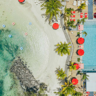 Luftaufnahme eines Resorts mit Sandstrand, klarem türkisfarbenem Wasser, Palmen und einem Schwimmbad mit roten Sonnenschirmen.