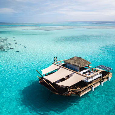 An overwater bar with a thatched roof in turquoise waters, featuring lounge areas with sail-like canopies and two people standing on the deck.