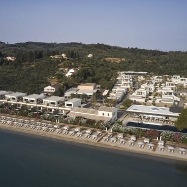 Luftaufnahme eines modernen Strandresorts mit weißen Gebäuden, Sonnenliegen und Sonnenschirmen entlang des Sandstrands, im Hintergrund grüne bewaldete Hügel.