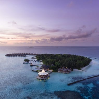 Luftaufnahme eines luxuriösen Inselresorts auf den Malediven während des Sonnenuntergangs, mit Wasserbungalows, üppigem Grün und umgebendem türkisem Wasser.