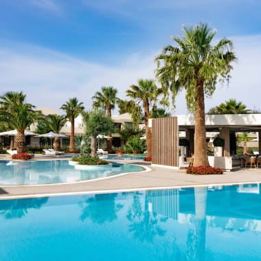 Luxuriöse Hotelpoolanlage mit Palmen, Sonnenschirmen und Sonnenliegen unter blauem Himmel.