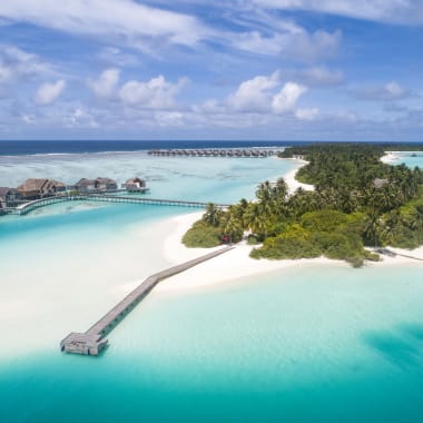 Eine tropische Insel mit üppigen Palmen und weißen Sandstränden, umgeben von klarem türkisfarbenem Wasser, mit hölzernen Stegen, die zu Villen führen, und einem langen Steg zur Insel.