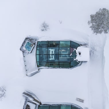 Aerial view of a snow-covered structure with a glass roof resembling an igloo, surrounded by snow and a few bare trees, with a hot tub on the left side and a bench nearby.