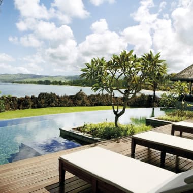 A luxurious outdoor pool area with lounge chairs, lush green trees, and a scenic view of water and mountains in the background.