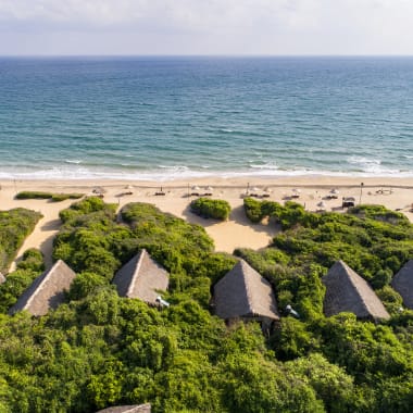 Luftaufnahme von stroh gedeckten Villen im Dschungel, umgeben von dichtem grünem Laub neben einem sandigen Strand, der zu einem ruhigen Ozean unter einem bewölkten Himmel führt.