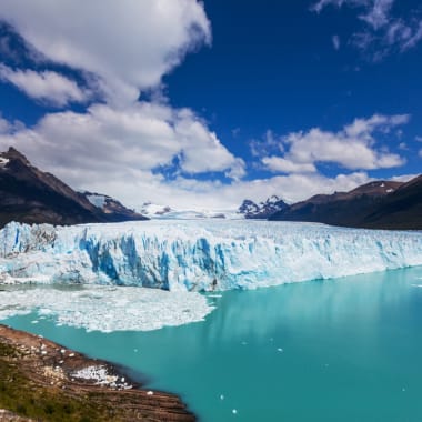 A massive glacier extending into a turquoise lake with mountains in the background under a partly cloudy sky.