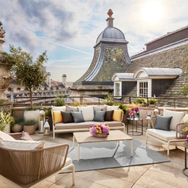 Ein stilvoller Dachterrassen-Lounge mit bequemen Sitzgelegenheiten, bunten Kissen und Blumenarrangements mit Blick auf historische Gebäude in London.