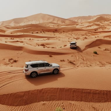 Zwei weiße SUVs fahren durch eine weite Wüstenlandschaft mit Sanddünen und Spuren im Sand