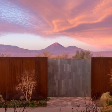 A modern outdoor gate with rust-colored metal panels, flanked by leafless trees and desert plants, with a mountain landscape and colorful sky in the background.
