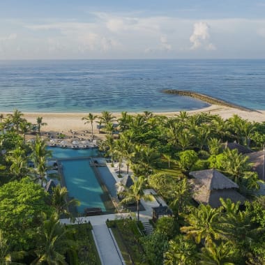 Luftaufnahme des Ritz-Carlton Bali Resorts mit üppigem Grün, Schwimmbädern, Wegen und dem Ozean im Hintergrund unter einem teilweise bewölkten Himmel.