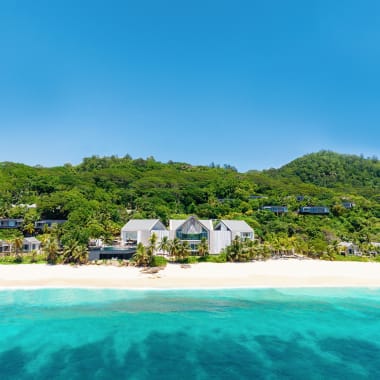 White modern beachfront villas nestled between a pristine sandy beach and lush green hills under a clear blue sky.