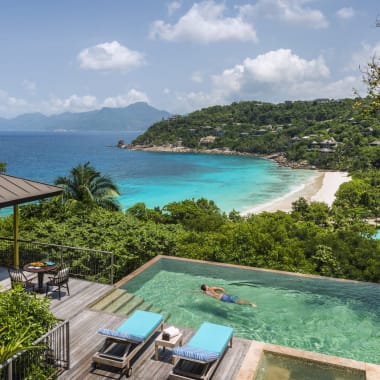 Eine Person schwimmt in einem Infinity-Pool einer luxuriösen Villa mit Blick auf einen tropischen Strand mit türkisfarbenem Wasser und üppigen grünen Hügeln im Hintergrund.