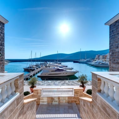 Ein malerischer Blick von einer Terrasse auf einen Yachthafen mit Booten in Montenegro, mit Bergen im Hintergrund unter einem hellen sonnigen Himmel.