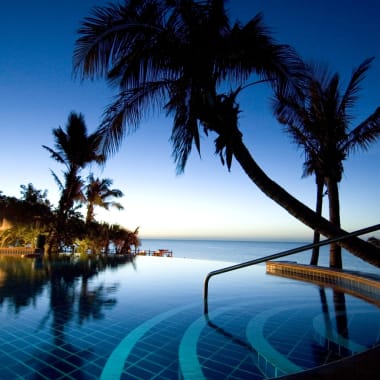 Nächtliche Sicht auf einen luxuriösen Resort-Pool, umgeben von Palmen auf einer tropischen Insel, mit dunklem Himmel und ruhigem Wasser.
