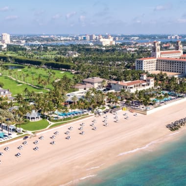 Aerial view of a luxurious beachfront resort with sun umbrellas and lounge chairs, alongside lush green gardens and a cityscape in the background.