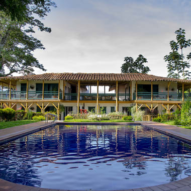 A luxurious two-story building with a large swimming pool in the foreground, surrounded by lush green trees and garden.