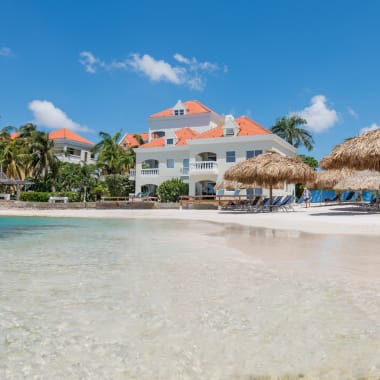 Ein luxuriöses Strandresort mit weißen Gebäuden und orangefarbenen Dächern, strandnahe Villen mit SchilfsonnenSchirmen und Liegestühlen am Sandstrand.