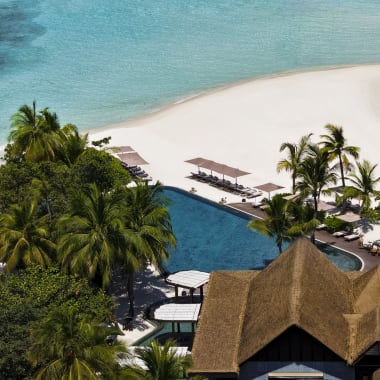 Luftaufnahme eines tropischen Inselresorts mit einem großen Schwimmbad umgeben von Palmen und strohgedeckten Gebäuden, angrenzend an eine weiße Sandbank, die in klares türkisfarbenes Wasser ragt.