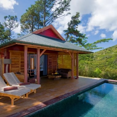 Eine hölzerne Lodge mit einem kleinen Pool in einer grünen, bergigen Landschaft, mit einem Outdoor-Sitzbereich und Liegestühlen.