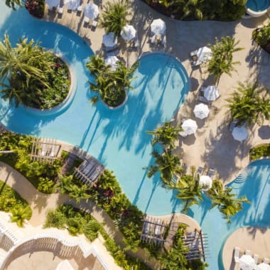 Eine Vogelperspektive auf einen Resort-Poolbereich mit mehreren kreisförmigen Pools, umgeben von Palmen, Liegestühlen und Wegen.