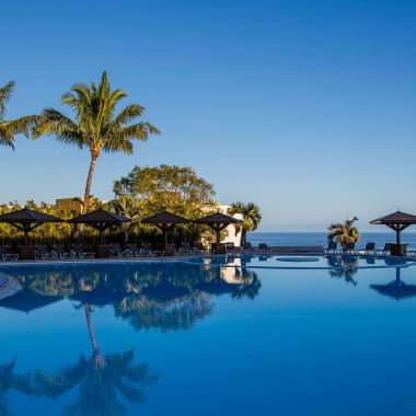 Unendlichkeits-Pool mit Palmen und Liegestühlen unter Sonnenschirmen mit Blick auf das Meer in einem tropischen Resort.