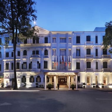 The exterior of the Sofitel Legend Metropole Hanoi hotel illuminated at dusk, with trees and a street in front.