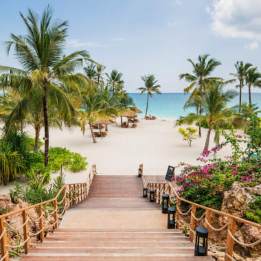 Wooden stairs leading down to a tropical beach with palm trees and turquoise water in the background.
