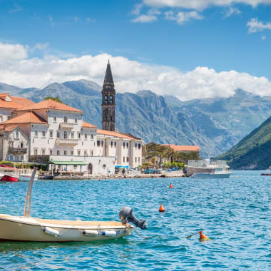 Ein malerischer Blick auf die Bucht von Kotor mit bunten Gebäuden, einem Kirchturm, einem Boot und Bergen im Hintergrund.