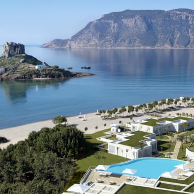 A scenic view of a luxury resort with swimming pools, modern white buildings, and a sandy beach along the coastline, with a rocky island and mountains in the background.