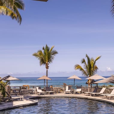 Ein geschwungener Swimmingpool umgeben von Palmen und Liegestühlen mit Sonnenschirmen, mit Blick auf das klare blaue Meer unter hellem Himmel.