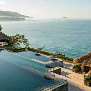 Ein mehrstufiger Infinity-Pool mit Blick auf den Ozean, mit Strohdachhütten in der Nähe und üppigem Grün im Hintergrund.