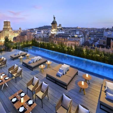Dachterrasse mit Liegestühlen, Tischen und einem Infinity-Pool mit Blick auf die Skyline von Barcelona bei Sonnenuntergang.