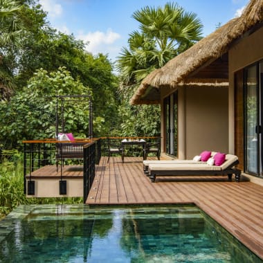 A luxurious outdoor patio with a small pool, lounge chair with cushions, and a thatched roof structure surrounded by lush green tropical trees.