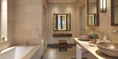 Luxuriöses Badezimmer mit beigen Stewwänden, Doppelwaschbecken mit Steinbeckenschalen und einer großen Badewanne, beleuchtet durch warmes Licht und eine dekorative Wandlampe.