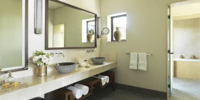 Luxuriöses Badezimmer mit Doppelwaschbecken, großen Spiegeln und natürlichem Licht durch Fenster, dekoriert mit Blumen und modernen Accessoires.