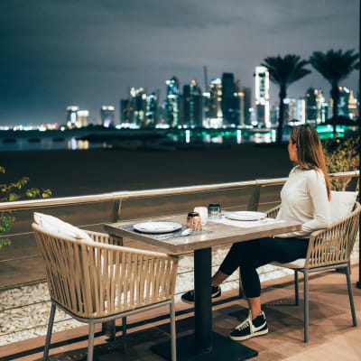 Eine Frau sitzt alleine an einem Tisch auf einer Außenterrasse mit Blick auf die beleuchtete Skyline von Doha bei Nacht.